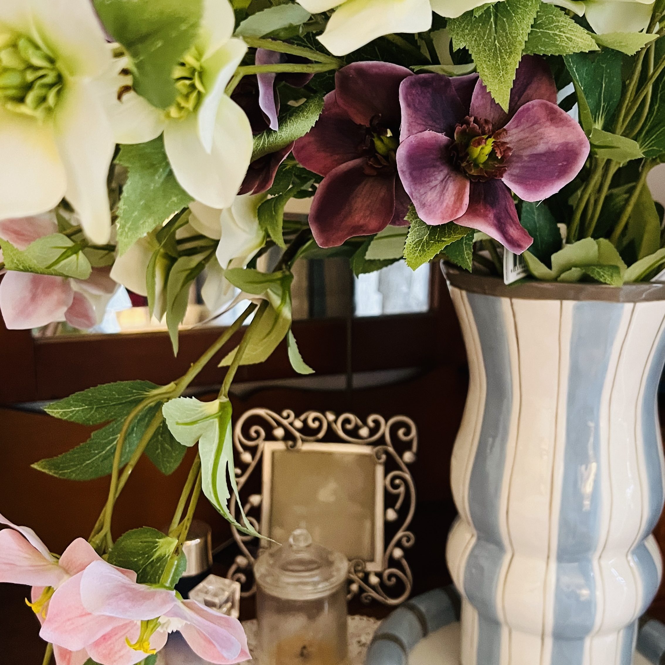 A decorative arrangement of flowers in a striped vase, featuring white, purple, and pink blooms, placed on a lace doily alongside a small glass jar and a picture frame.